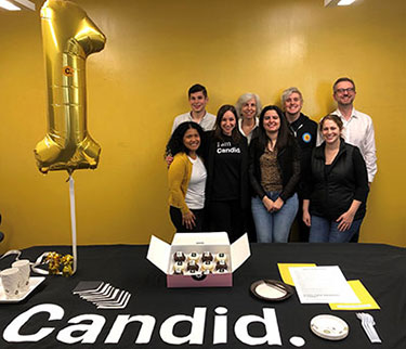 Three men and five women from Candid's D.C. office celebrate Candid's first anniversary with cupcakes and a balloon.
